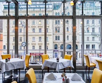 Bright commercial restaurant interior with dining tables, yellow chairs and large city-facing windows.