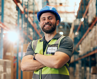 Warehouse worker wearing a hard hat and safety vest in an industrial storage facility.