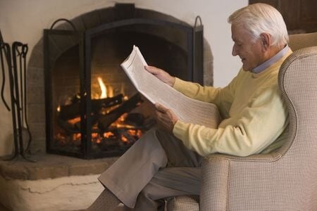 Elderly man in room with fireplace reading a newspaper